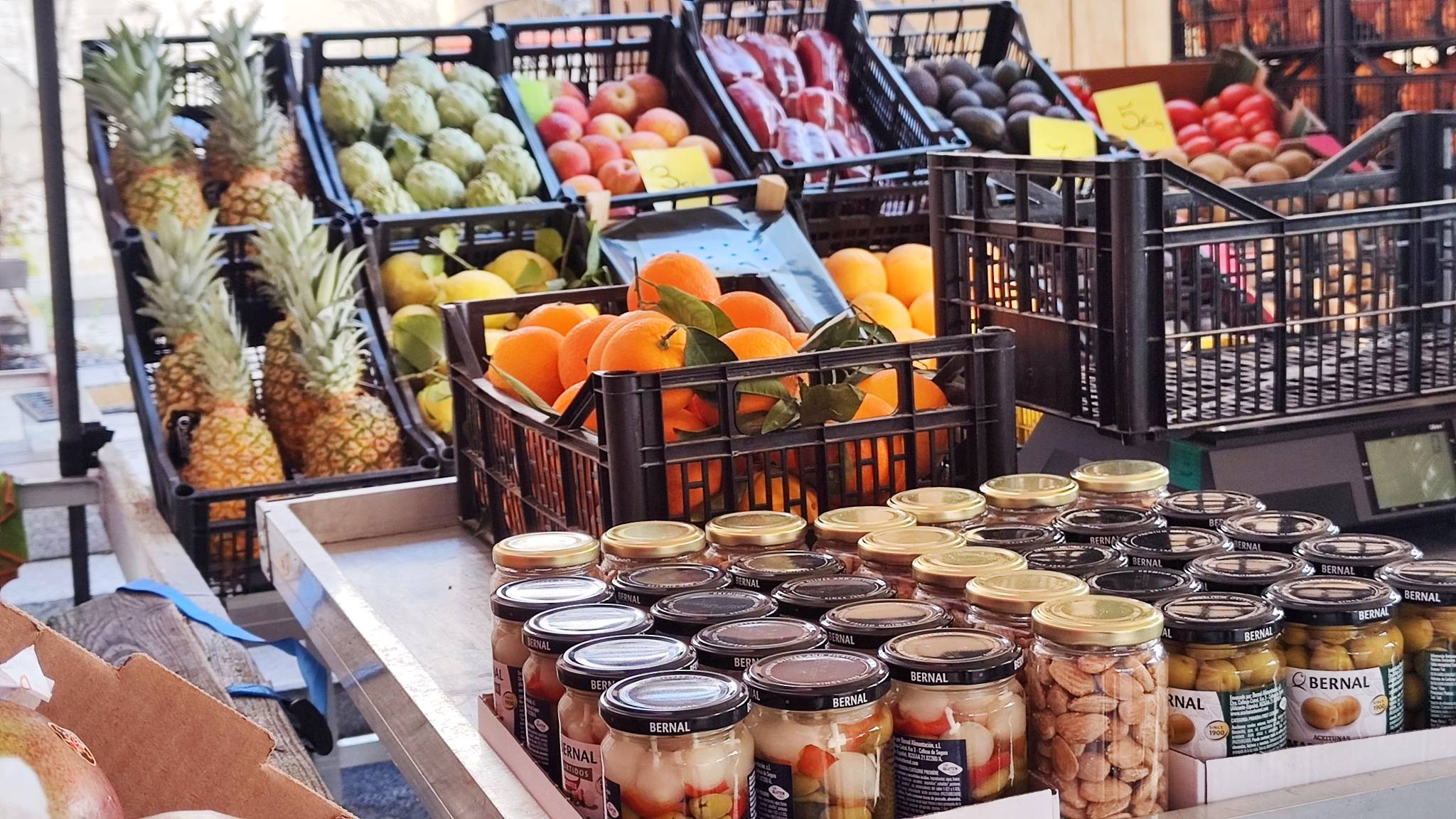 A table with various fruits and jars of food in black crates and cardboard boxes.