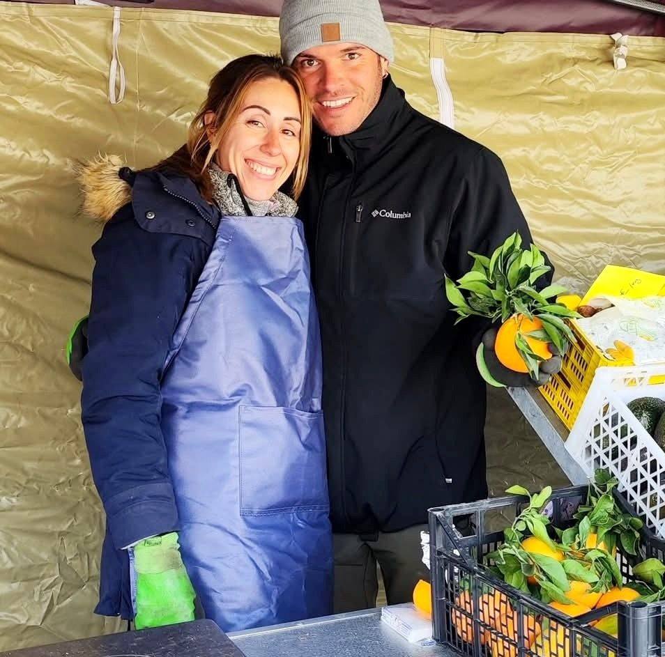 A man and woman, dressed in winter attire, stand together in a tent, surrounded by crates of oranges and leaves. The woman...